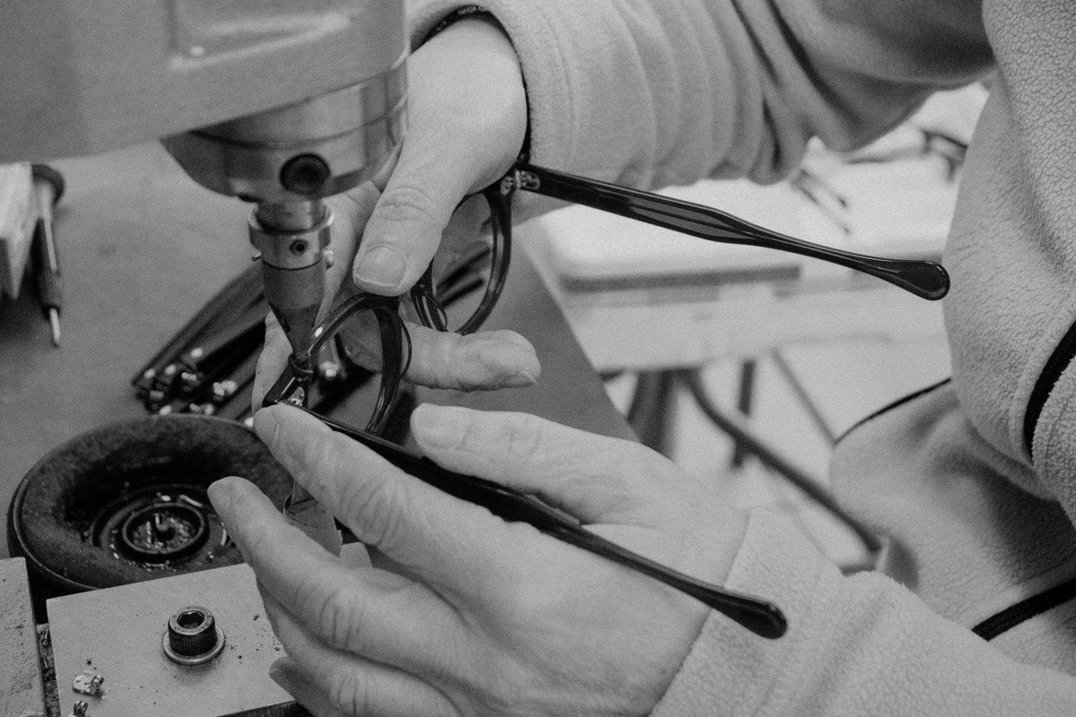 Black-and-white close-up of a craftsperson working on a spectacle frame at a workshop machine.