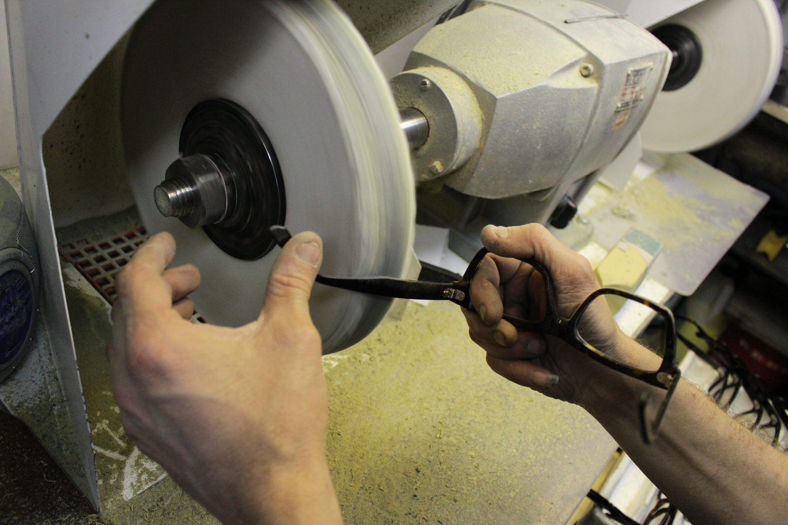 Craftsperson polishing a spectacle arm by hand against a rotating buffing wheel.