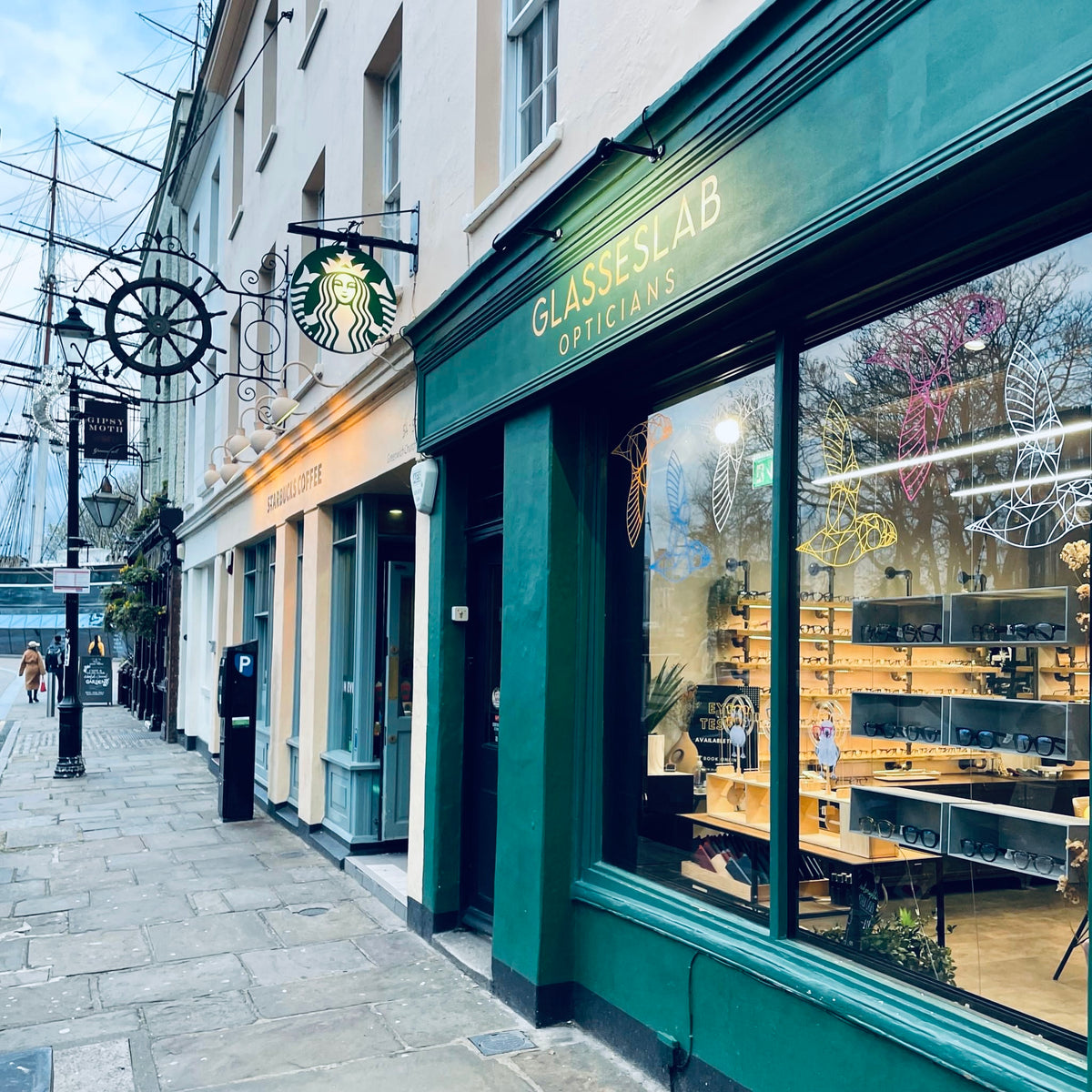 Green Glasseslab Opticians shopfront with illuminated eyewear displays beside a pedestrian street.